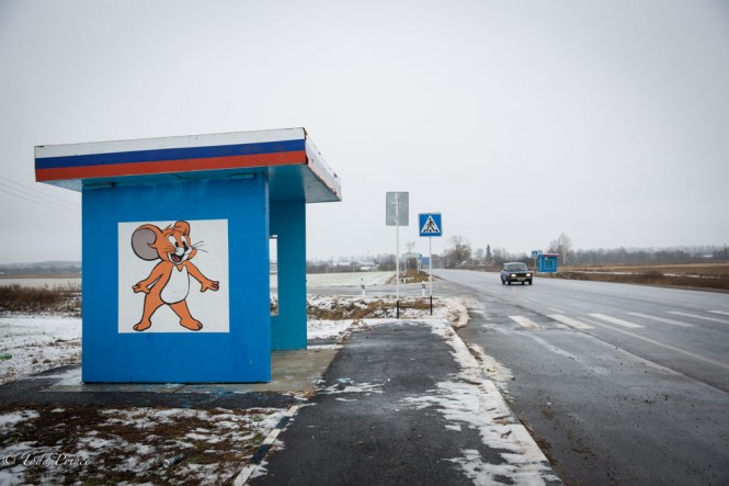 A Russian lada drives down a kursk highway, passing a bus stop with Jerry the cartoon character painted on it.