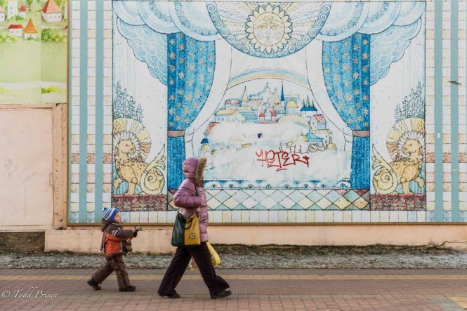 A mother and child walking past a Ramenskoe city wall covered in a village scene. 