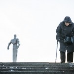 An elderly woman walks down stairs as Gagarin statue rises behind her.