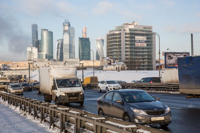Cars pass along a major city road as Moscow's Financial District basks in the sunlight in the background.